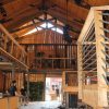 Interior of a wooden barn with high ceilings and metal railings, featuring a second-level balcony and light fixtures. Two people are present, and plants are visible in the background.