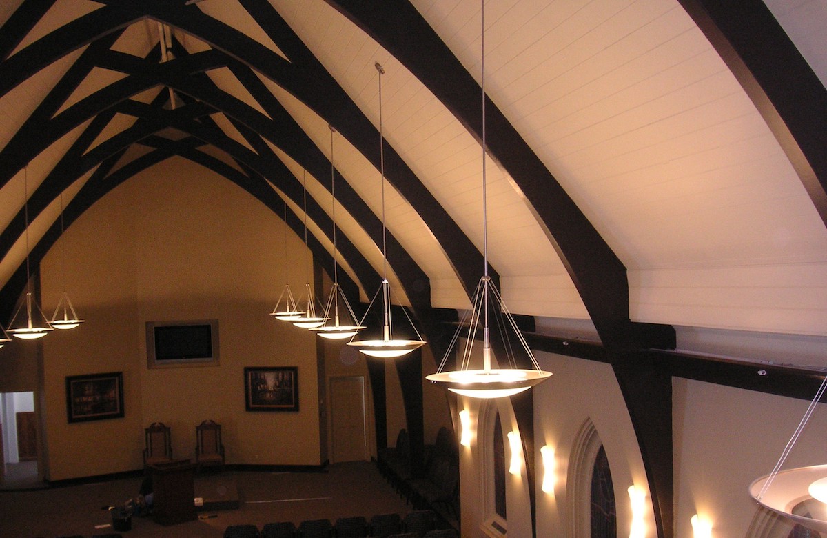 A church interior with a high, vaulted ceiling, dark wooden beams, and several pendant lights hanging down