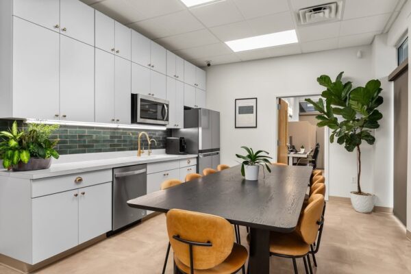 Modern veterinary staff break room with a long dining table, tan chairs, white cabinetry, and plants.