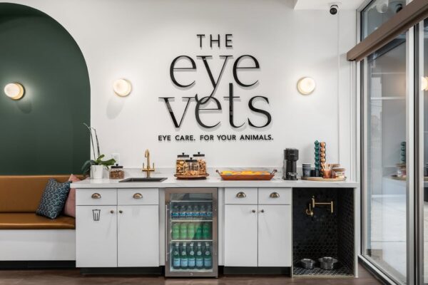 Client refreshment station at a veterinary clinic, featuring a beverage fridge, coffee maker, and pet water station under a wall sign for The Eye Vets.