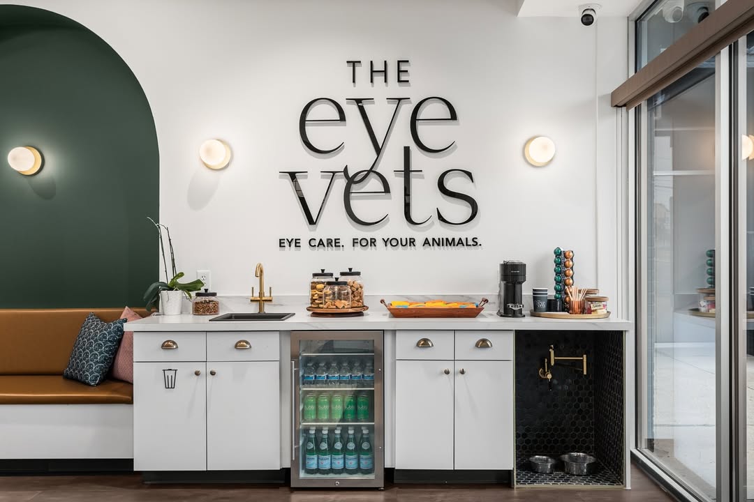 Client refreshment station at a veterinary clinic, featuring a beverage fridge, coffee maker, and pet water station under a wall sign for The Eye Vets.